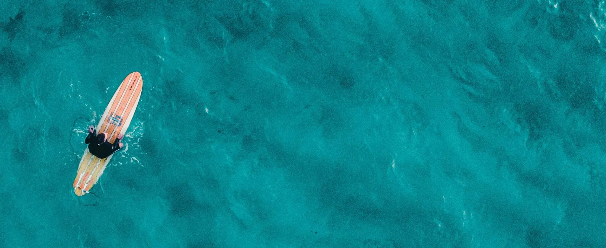 A solitary surfer on a longboard in vast turquoise ocean, seen from above.