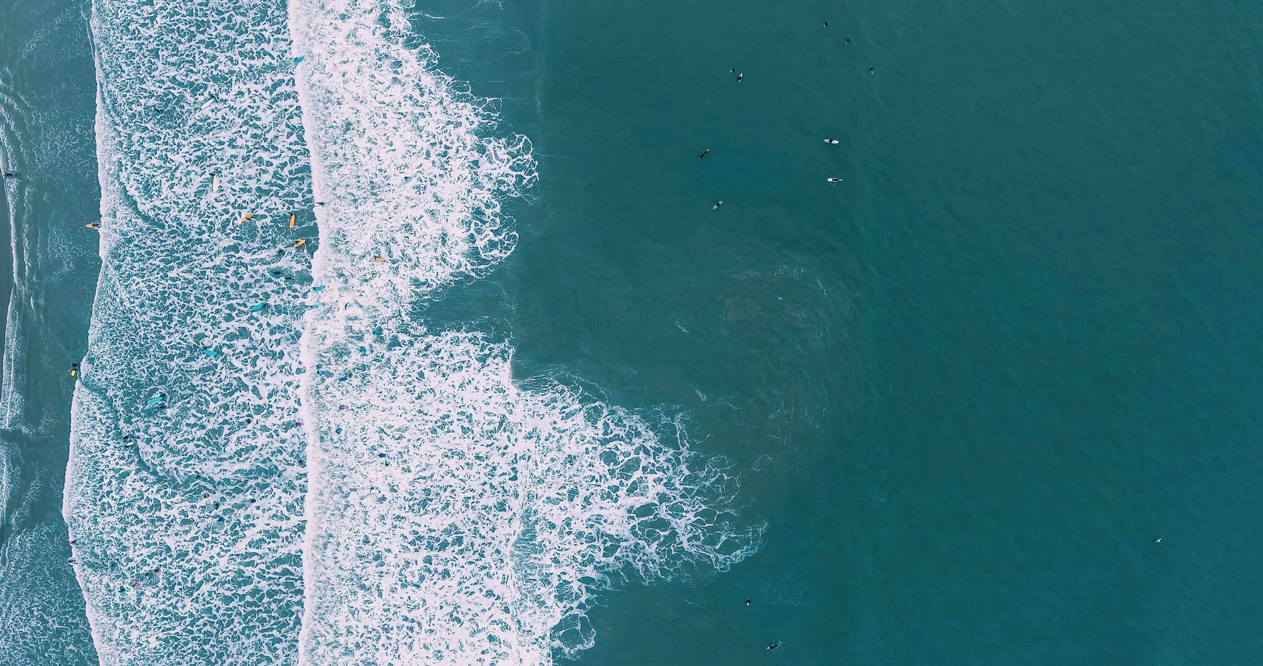 Aerial view of surfers in breaking surf, with white foam patterns against deep teal water.