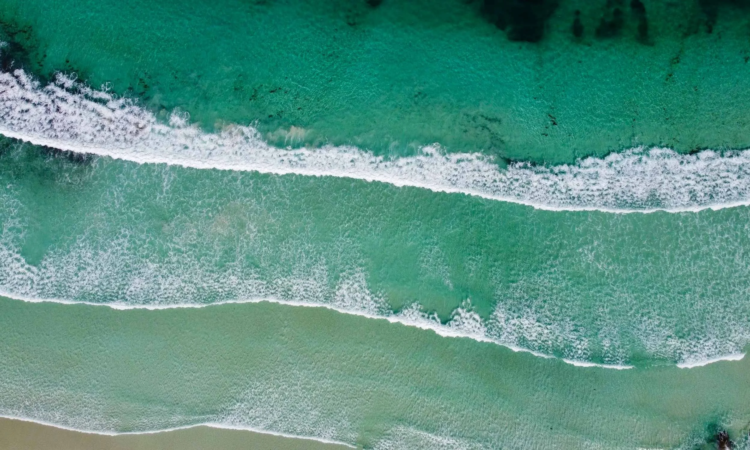 Aerial view of a beach where teal ocean meets pale sand in a horizontal band of white surf.