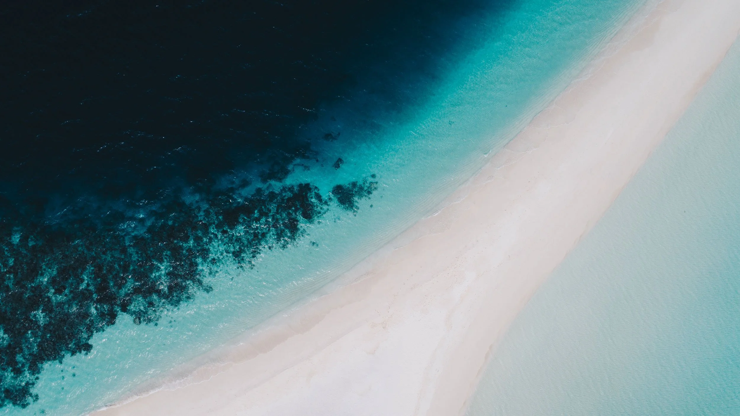 Aerial view of a coastline where deep teal water meets white sand.