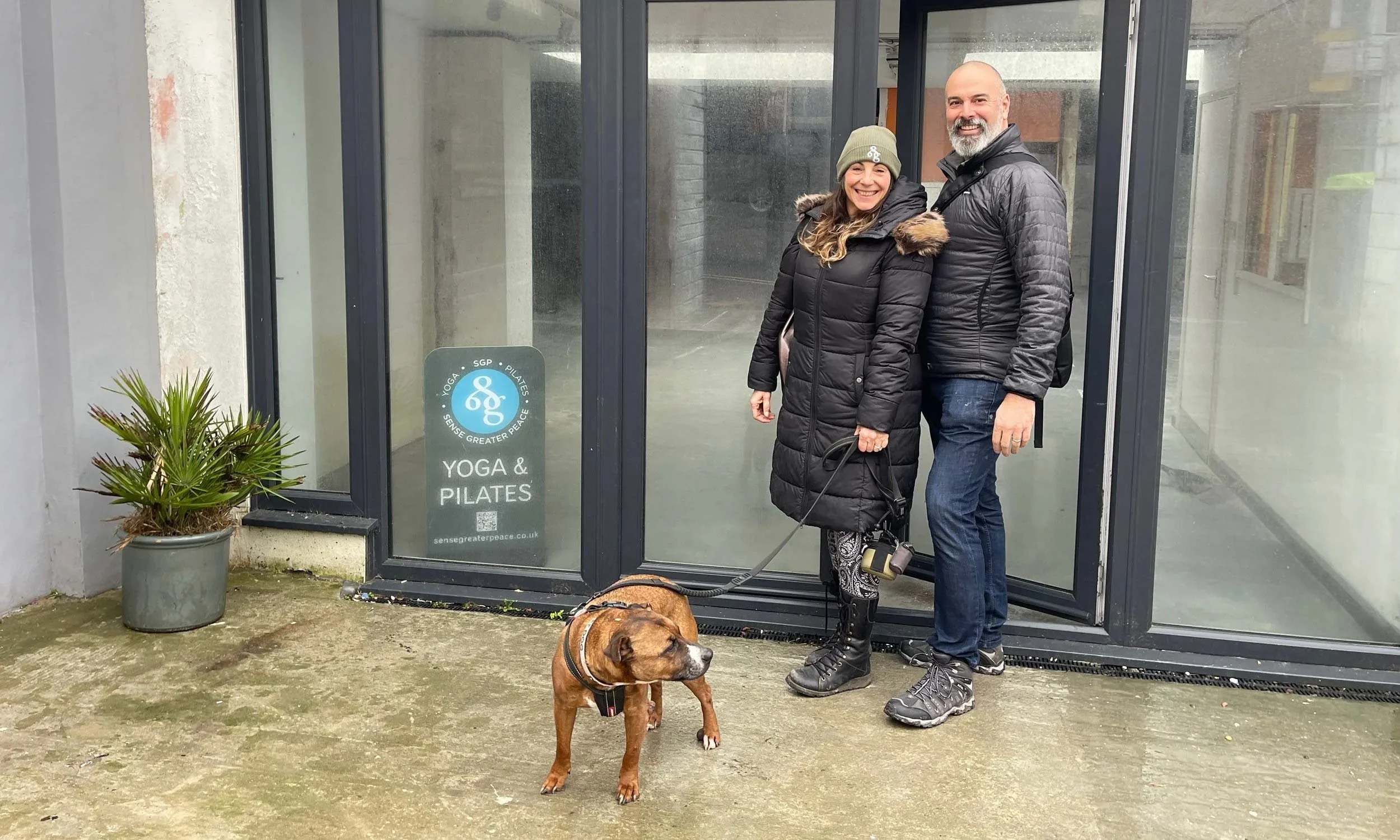 A Peter Dawson and Sue Dawson and their dog stand outside a studio entrance signed Sense Greater Peace and Yoga and Pilates.