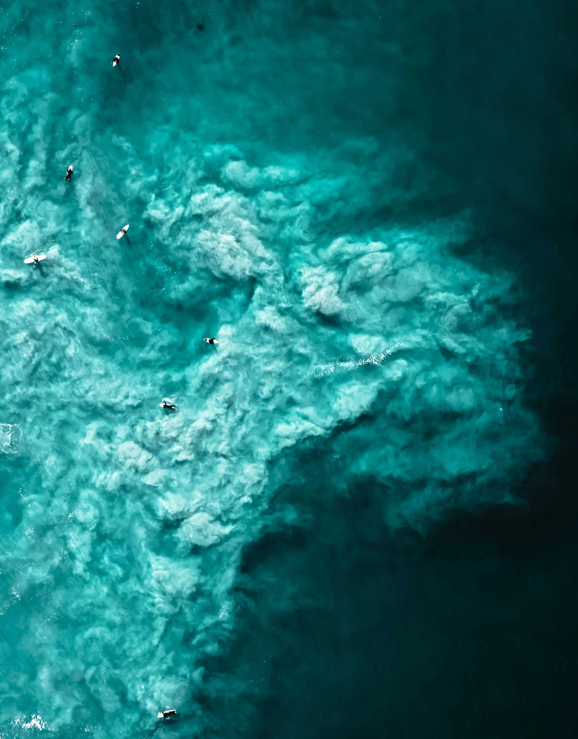 Aerial view of surfers waiting in turquoise ocean with swirling white-water patterns.