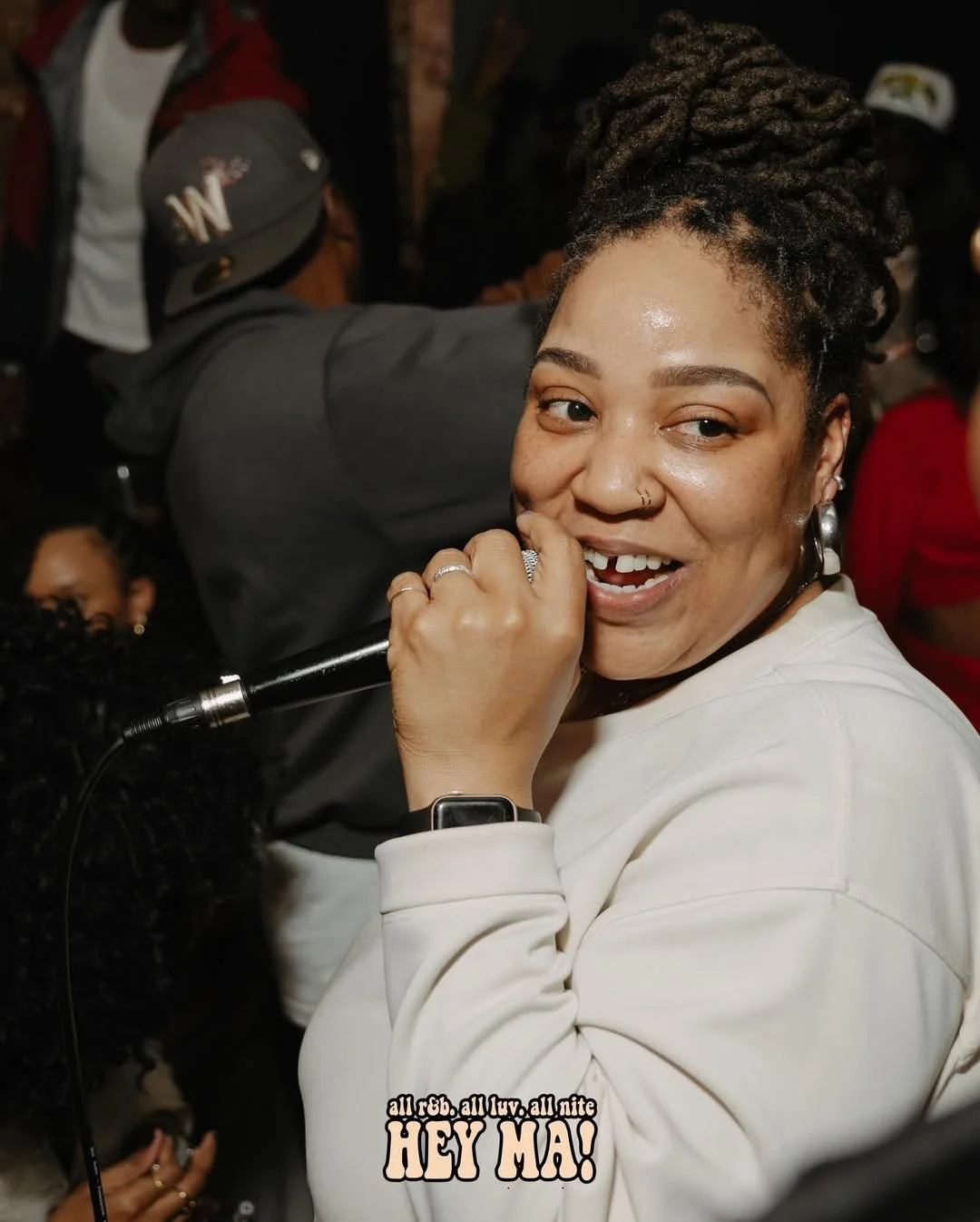 A woman with braided hair and hoop earrings holding a microphone, smiling at the camera in a crowd at a party or concert.