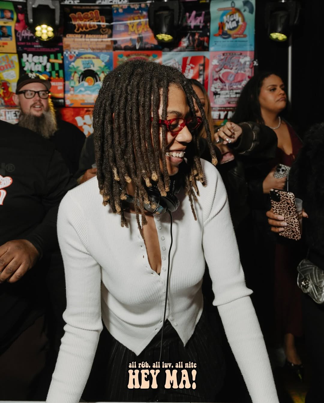 A woman with dreadlocks, red glasses, and a white sweater smiling at a lively party or event, with colorful posters on the wall behind and other people holding drinks around her.