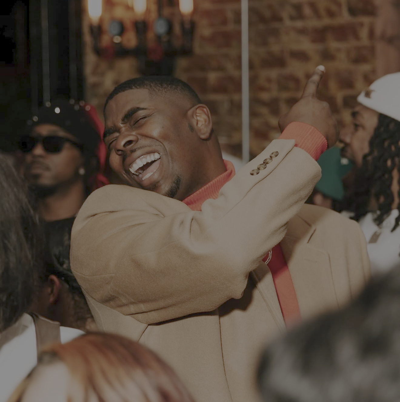 A man in a beige jacket and red shirt has his eyes closed and is smiling, dancing at a party with other people around him.