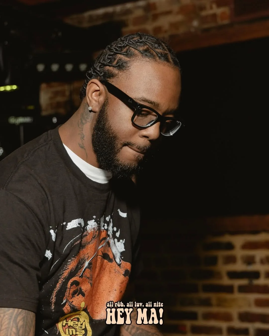 A young man with braided hair, glasses, and a beard, wearing a graphic T-shirt indoors with a brick wall background, looking down thoughtfully.