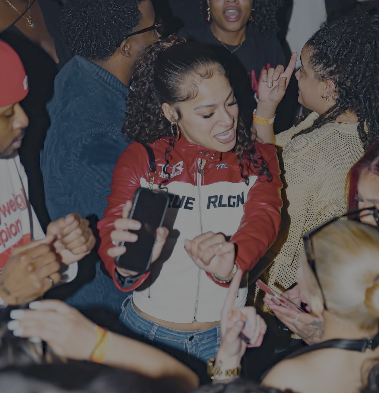 Group of people surrounding a woman with curly hair in a red and white jacket, holding a smartphone, smiling and engaging in conversation.