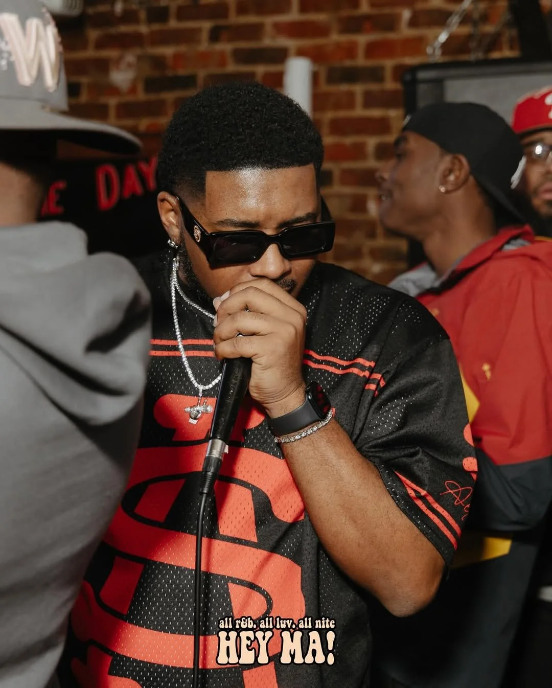 Young man wearing sunglasses, jewelry, and a black and red sports jersey holding a microphone, with a brick wall background and others in casual clothing nearby.