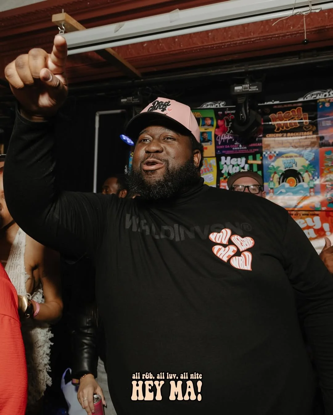 A man with a beard is dancing and smiling at a party, wearing a pink cap and a black shirt with colorful print, surrounded by other people and colorful event posters in the background.