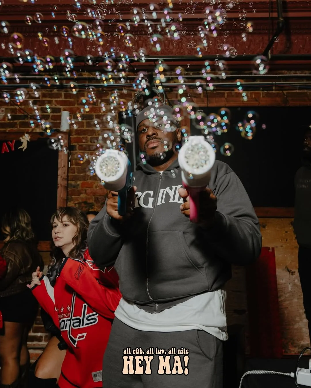A man in a gray hoodie and gray sweatpants holding two bubble guns, with bubbles floating in the air, in a social setting with a brick wall background and other people around.