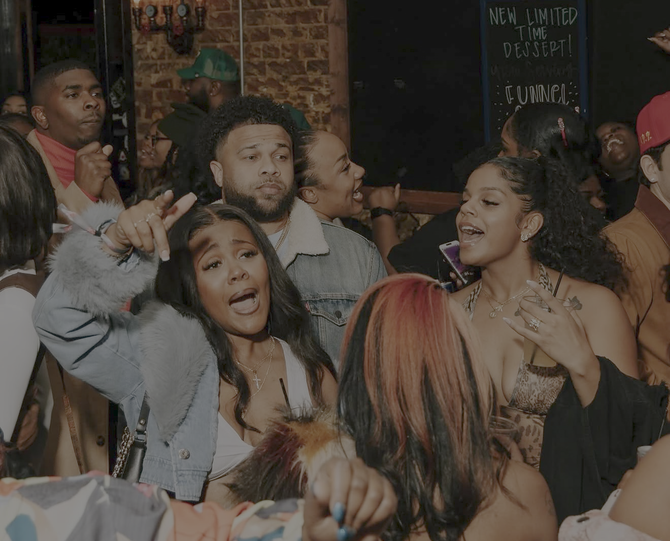 A group of diverse people gathered at a social event, engaging in conversation and showing varied expressions of excitement and joy. The background features a brick wall and a sign promoting a new dessert.