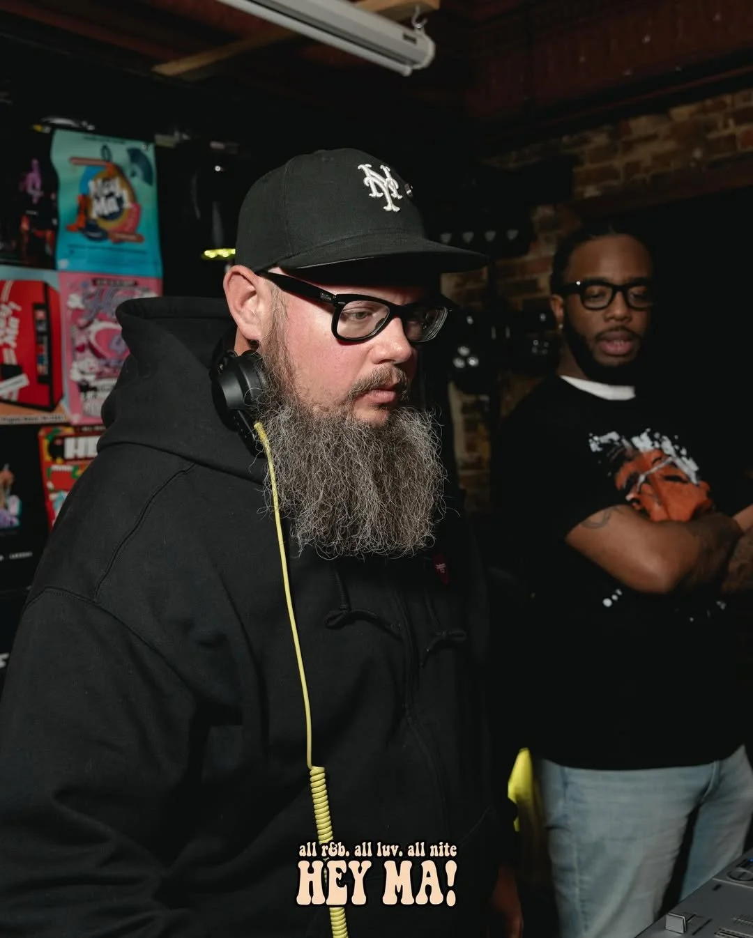 A man with glasses, a black cap, and a long beard wearing a black hoodie and yellow headphones, standing next to another man with glasses and a tattoo on his arm, in a room with arcade games and exposed brick walls.