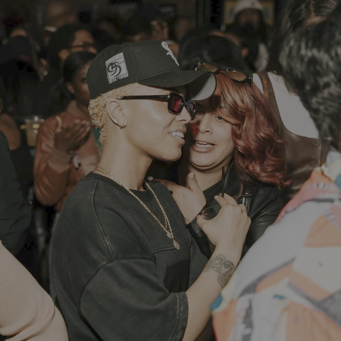 Two women with close hairstyles and sunglasses leaning in and smiling, in a crowded social gathering.