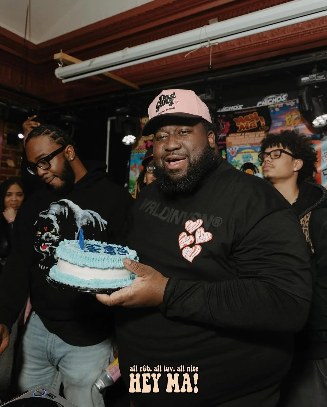 A man holding a birthday cake with a blue candle, surrounded by friends at a celebration, with colorful posters in the background.