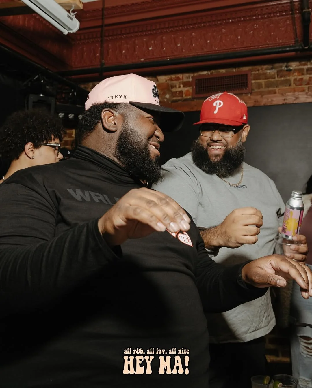 Two men smiling and enjoying drinks at a social gathering in a bar or restaurant, with a brick wall and ceiling air conditioning unit in the background.