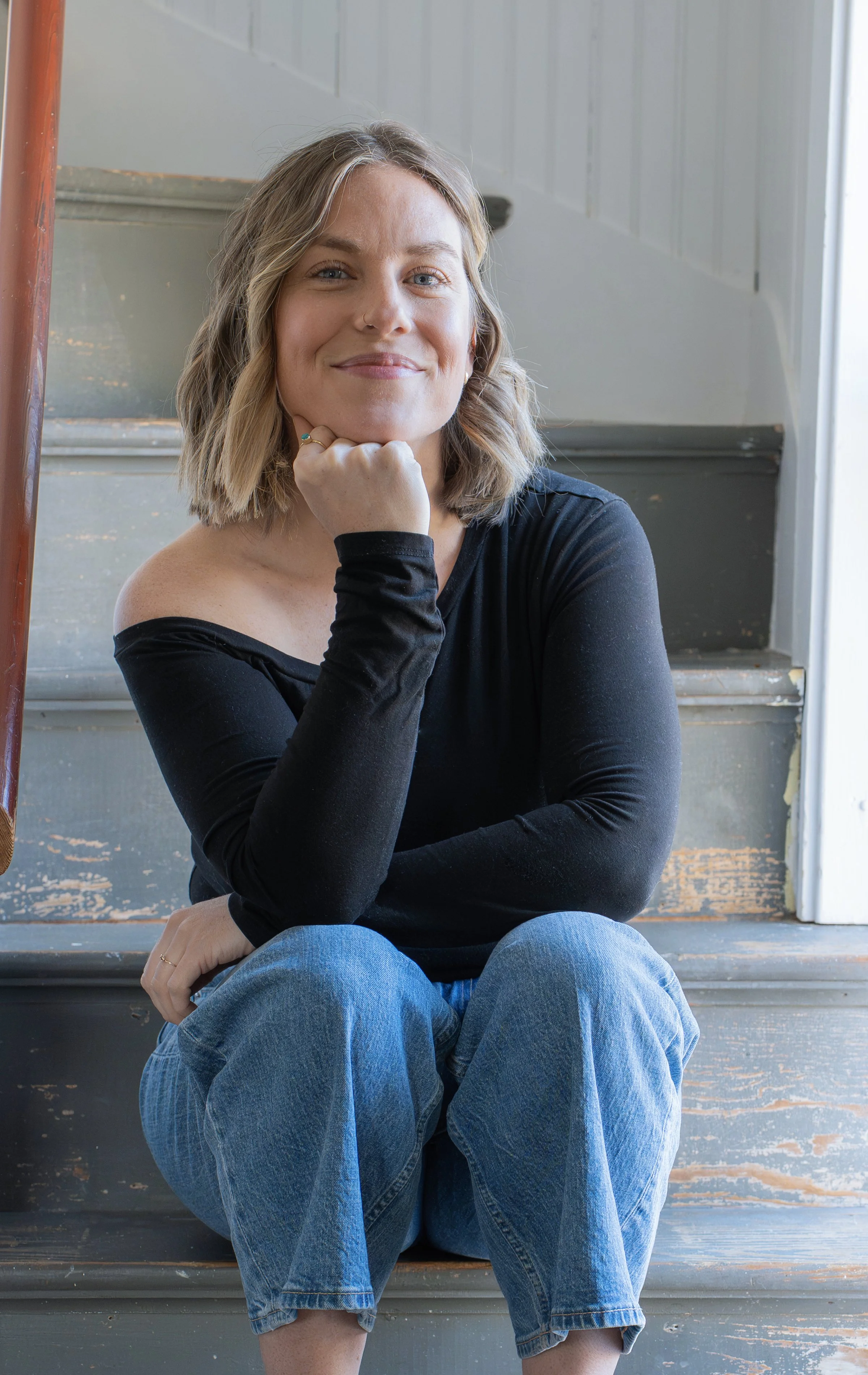 A woman sitting on stairs with her chin resting on her hand, smiling. She has blonde, shoulder-length hair, wearing a black off-shoulder top and blue jeans.