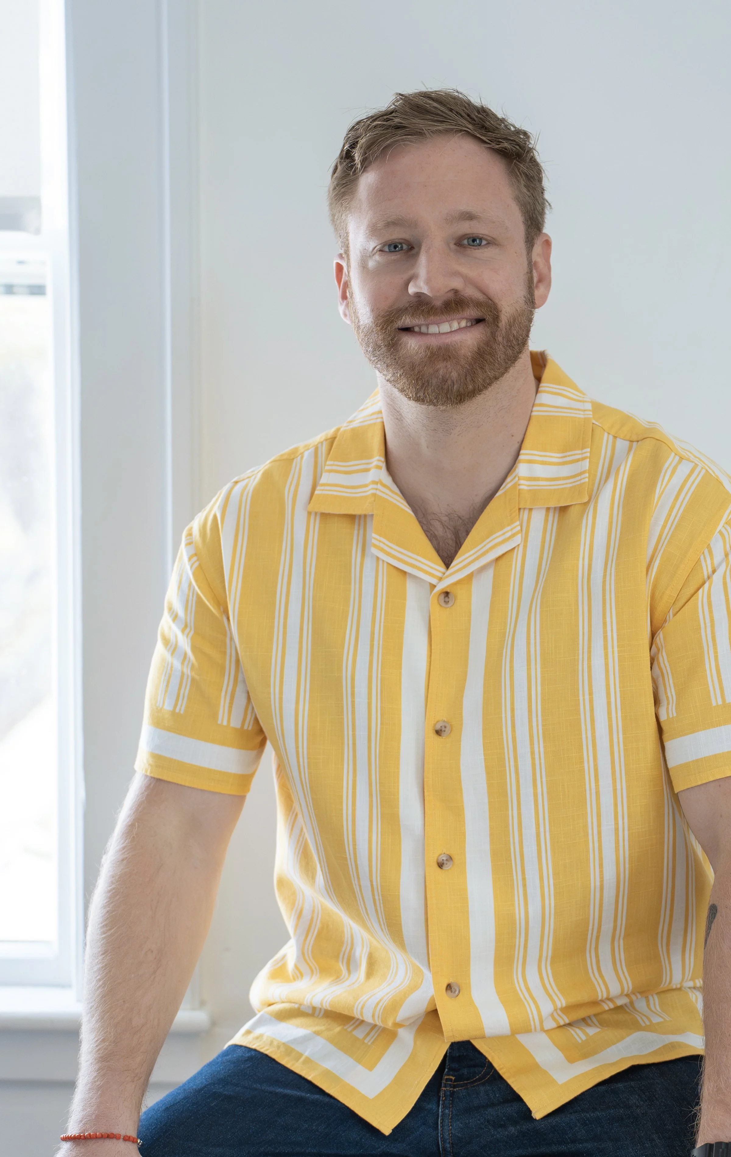 A man with a beard and short hair smiling, wearing a yellow and white striped shirt, sitting indoors near a window.