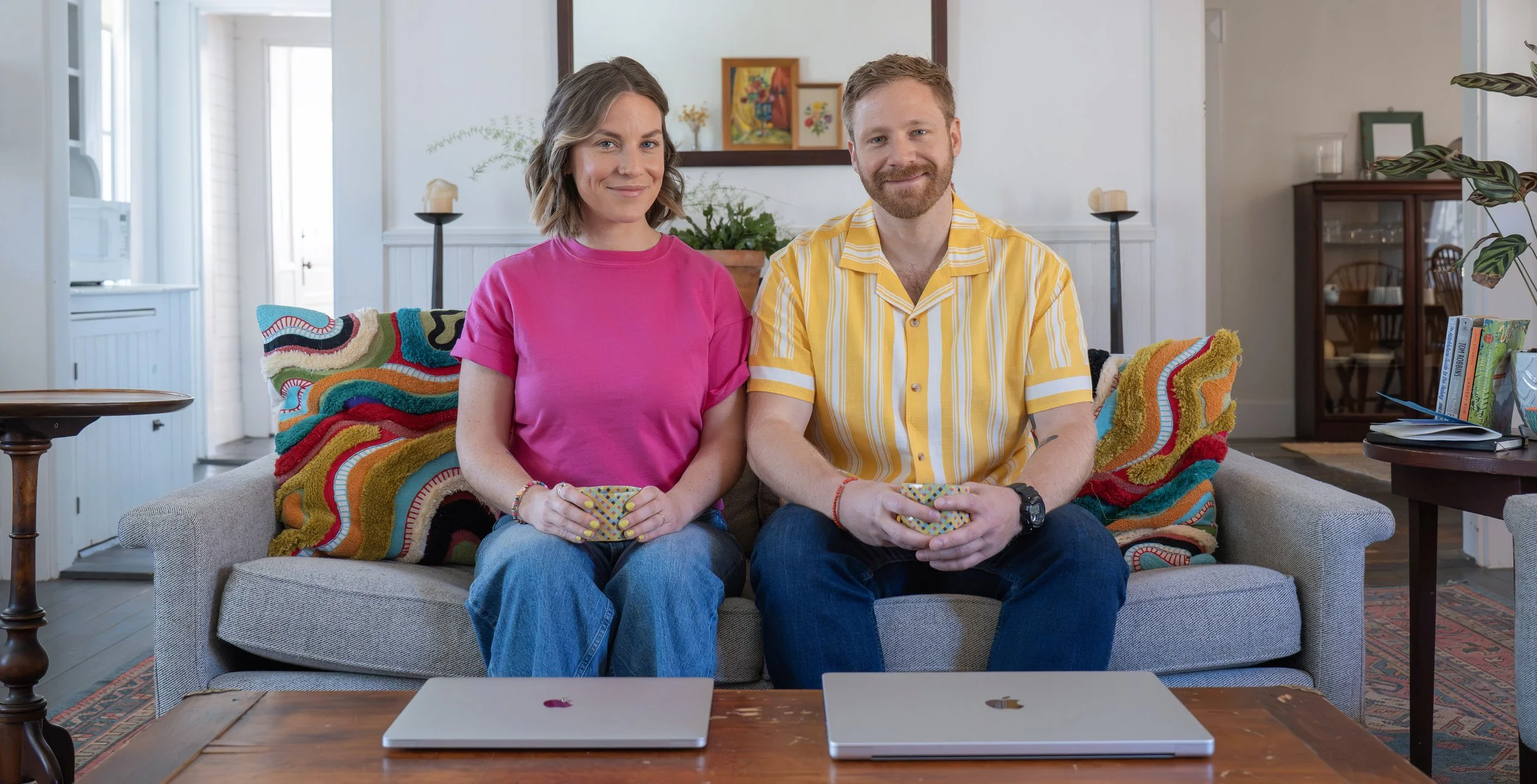 A smiling woman with shoulder-length hair wearing a pink t-shirt and a man with a beard wearing a yellow striped shirt sitting on a colorful, patterned couch holding cups, in a cozy living room with books and plants.