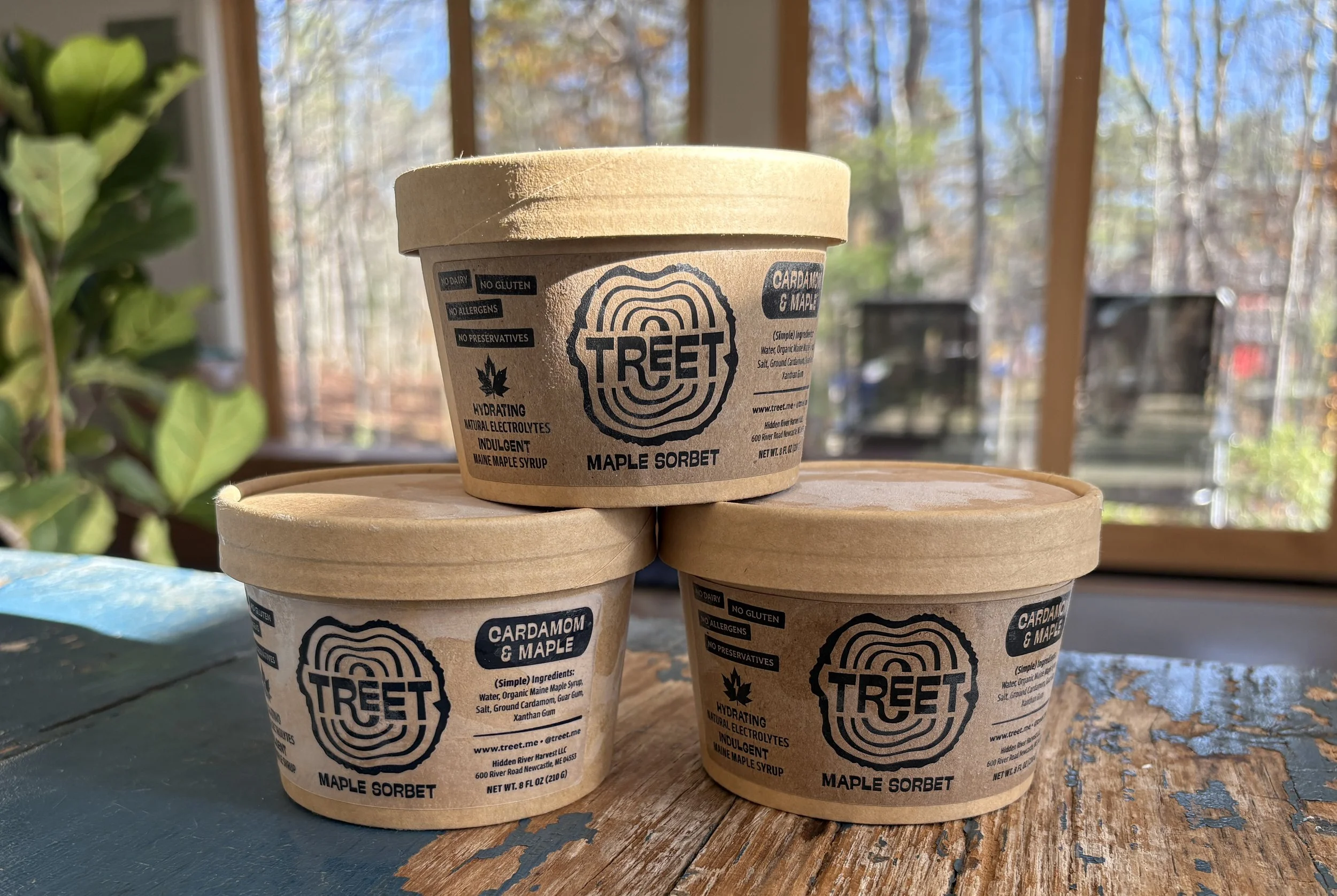 Several containers of Treet maple sorbet stacked on a wooden surface inside a room with large windows showing trees and blue sky outside.