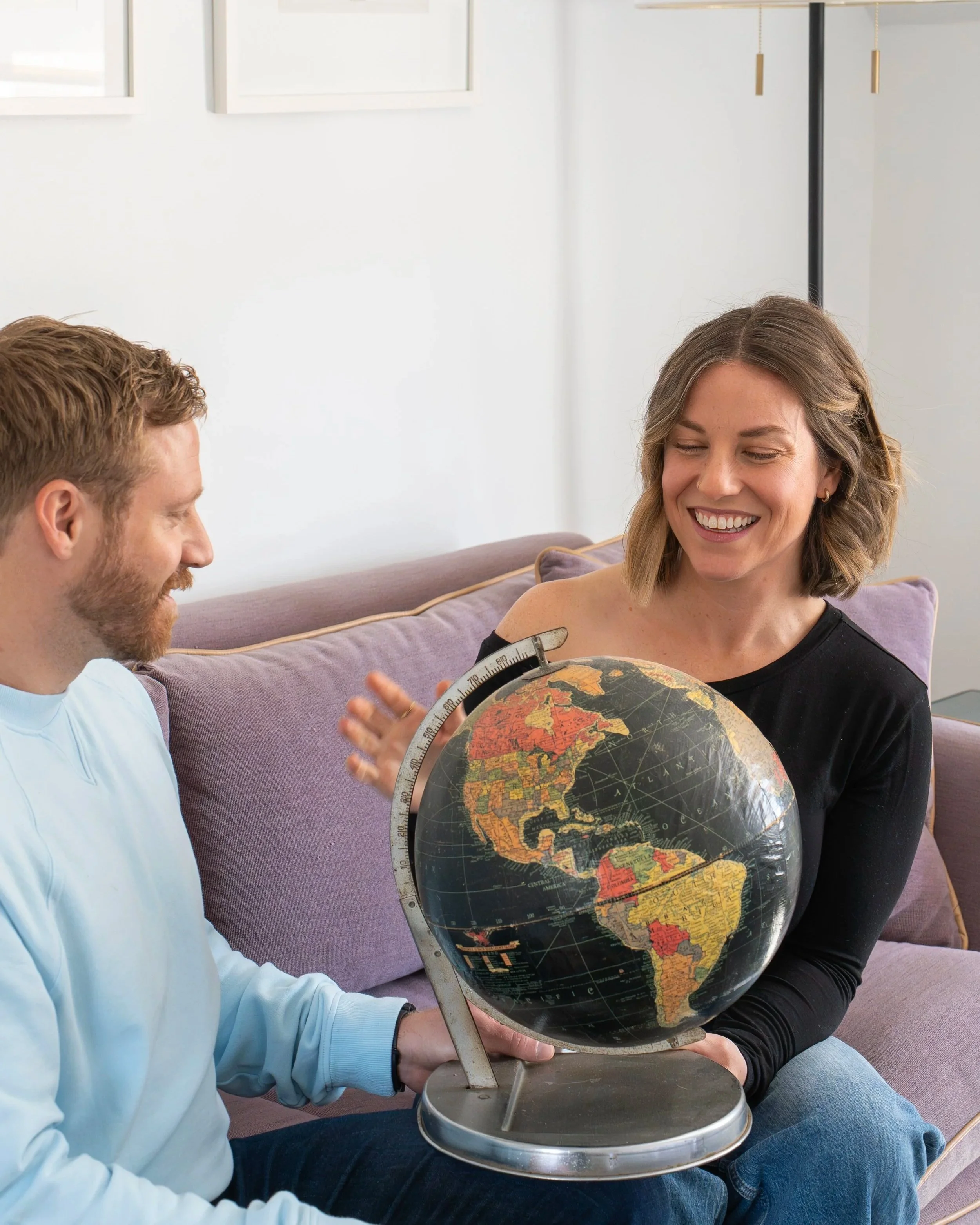 Two people sitting on a couch smiling while looking at a globe. The woman is holding the globe, and the man appears to be engaged in the conversation.