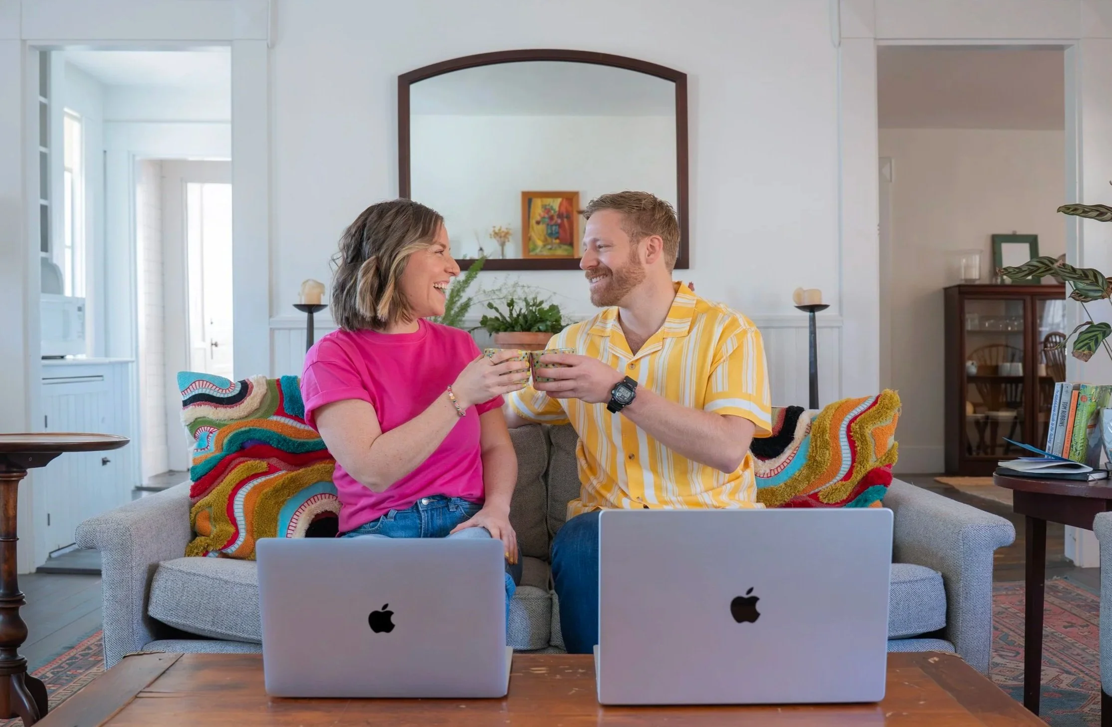 A smiling woman and man toasting with mugs in a living room with two open laptops on a table in front of them. They are sitting on a sofa with a colorful, patterned blanket. The room features a large mirror, potted plants, books, and a wooden cabinet.