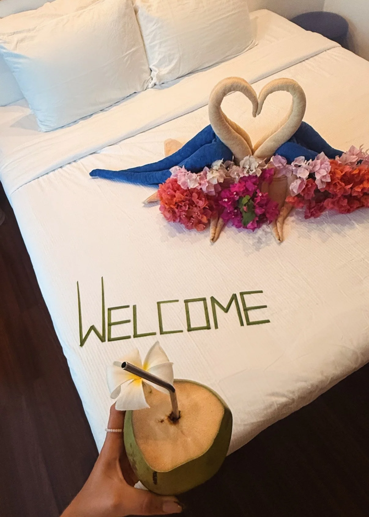 Une décoration de lit avec des serviettes en forme de cœur et de cygnes, des fleurs colorées, un message 'WELCOME' écrit avec des feuilles vertes, et une boisson dans une noix de coco avec une fleur blanche