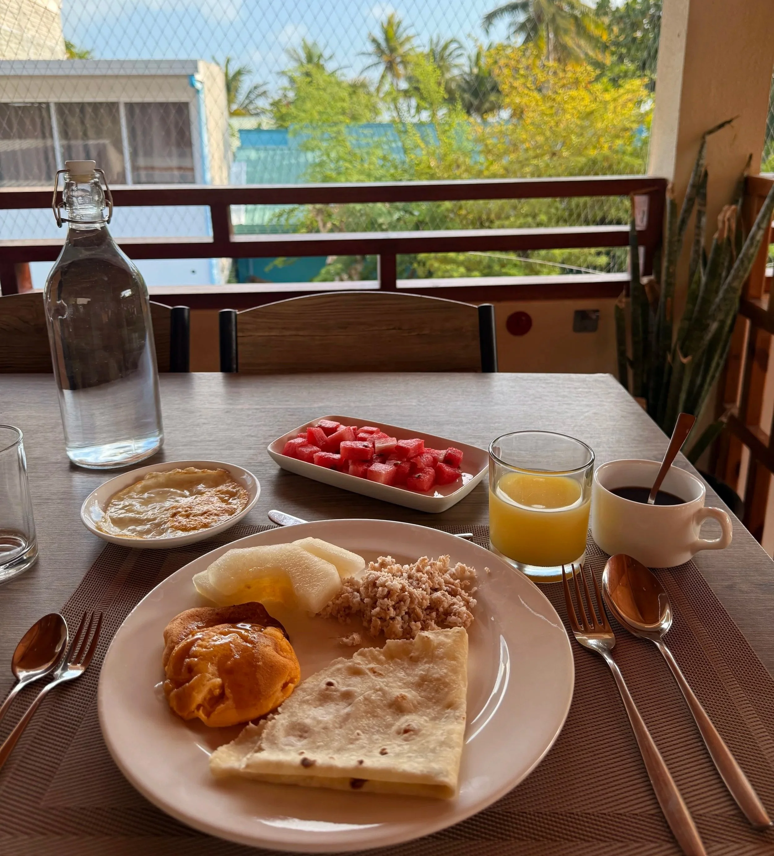 Petit déjeuner avec des œufs, du pain, des crêpes, de la crème ou du fromage, des fruits, un verre de jus d'orange, une tasse de café ou de thé, une bouteille d'eau en plastique, et une assiette avec des morceaux de pastèque, posé sur une table en bo