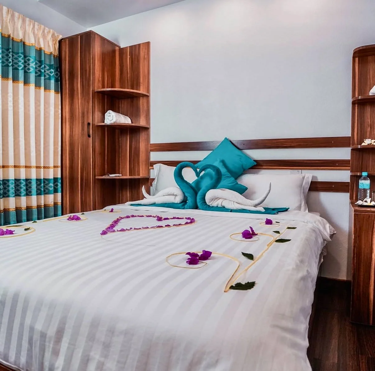 Lits dans une chambre d'hôtel décoré avec des serviettes en forme de cygnes et des fleurs, avec des coussins bleus, un comptoir en bois, une bouteille d'eau, et des rideaux colorés.