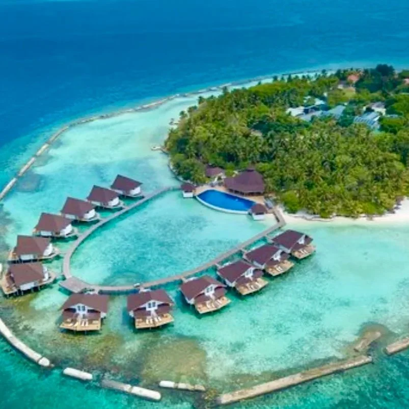 Vue aérienne d'un complexe hôtelier avec des villas sur pilotis entourées d'eau turquoise, en face d'une île verte avec des bâtiments et une piscine.