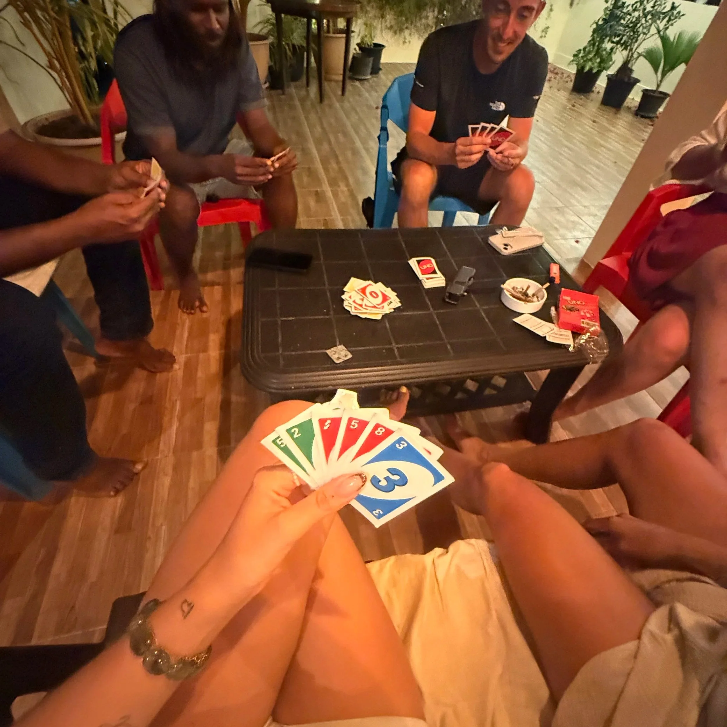 Les personnes jouent au Uno, avec des cartes rouge et jaune sur une table noire, dans une pièce décorée avec des plantes vertes et un sol en bois. L'ambiance est détendue et amicale.