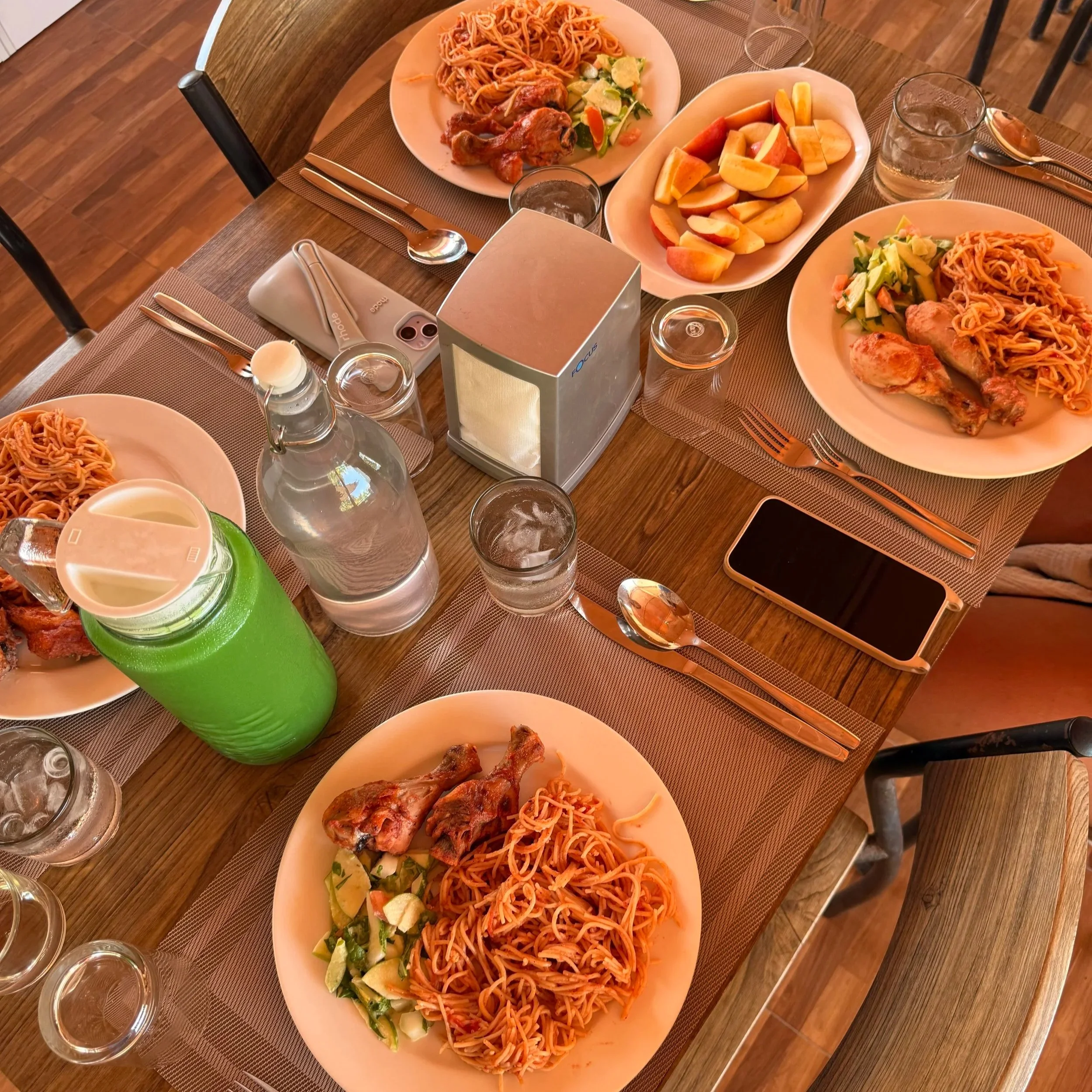 Assiette de spaghetti avec morceaux de poulet et salade de légumes, accompagnée de pommes et de verres d'eau, sur une table en bois.