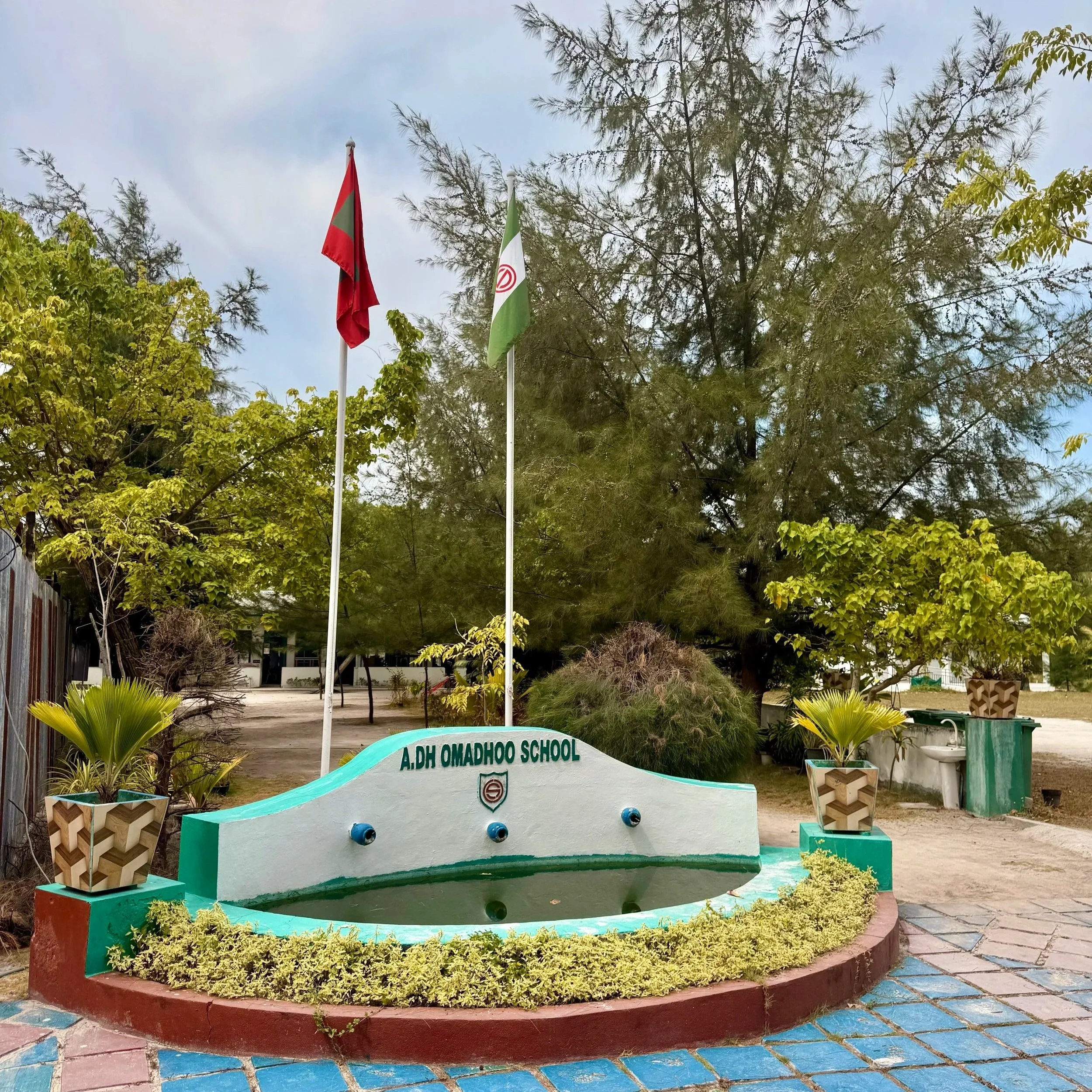 Fontaine devant une école nommée A.DH Omadhoo School avec des arbres et un ciel nuageux en arrière-plan.