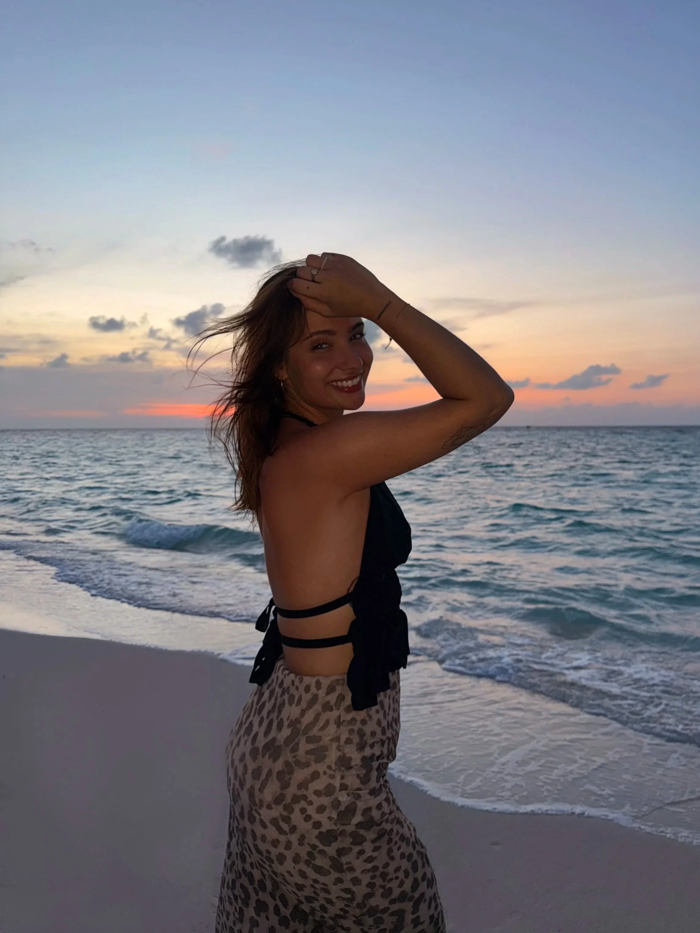 Une femme souriante devant la mer au coucher du soleil, portant un haut noir et une jupe à motif léopard, levant une main dans ses cheveux.