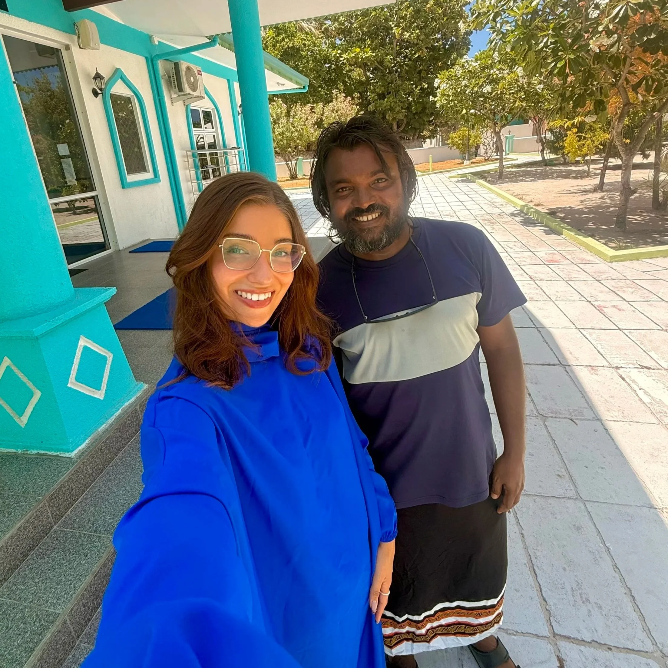 Deux personnes prennent un selfie devant un bâtiment peint en blanc et turquoise, avec fenêtres et climatiseurs, entouré d'arbres.