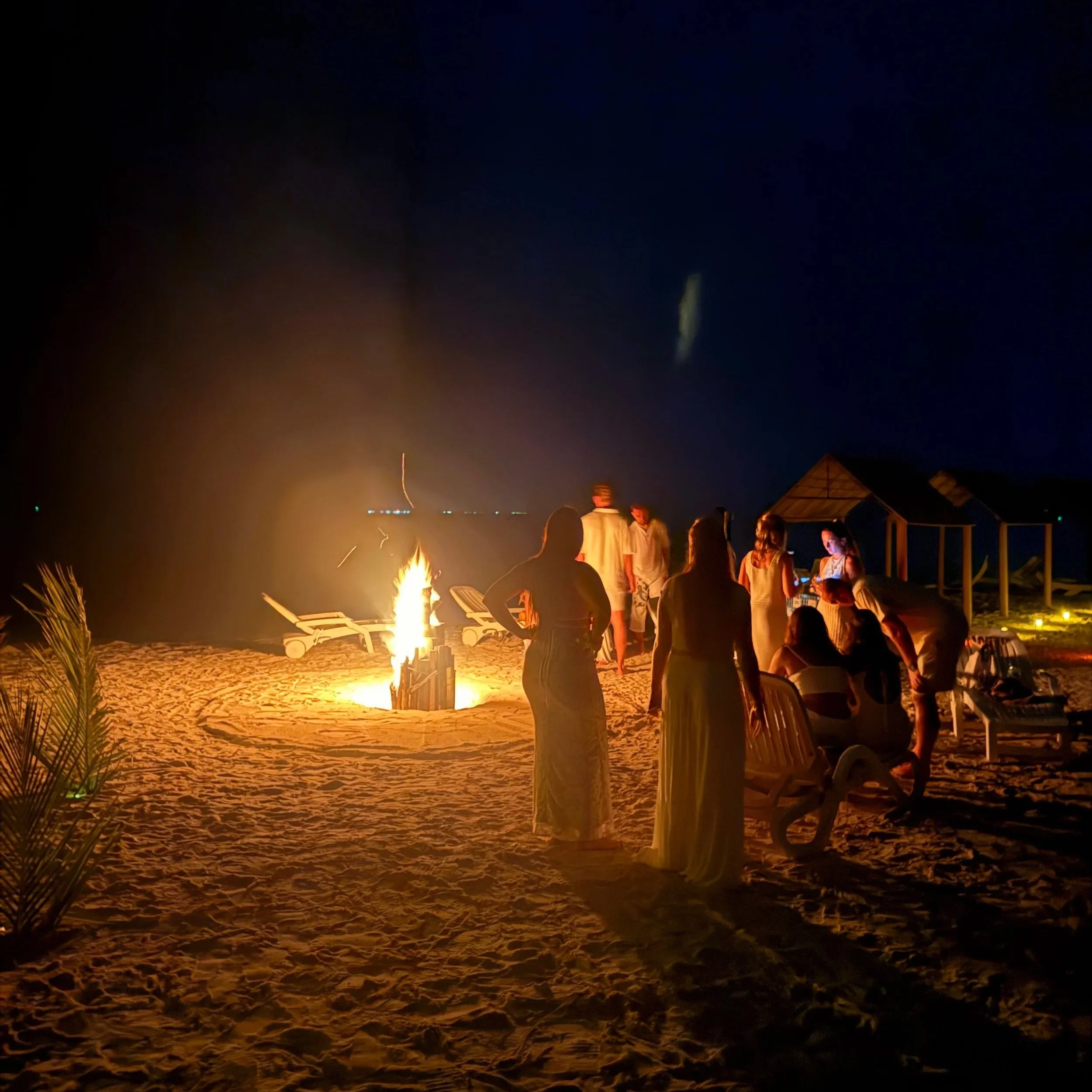 Fête sur la plage la nuit avec un feu de camp, des personnes assises sur des chaises et discutant, un bâtiment, et un ciel étoilé