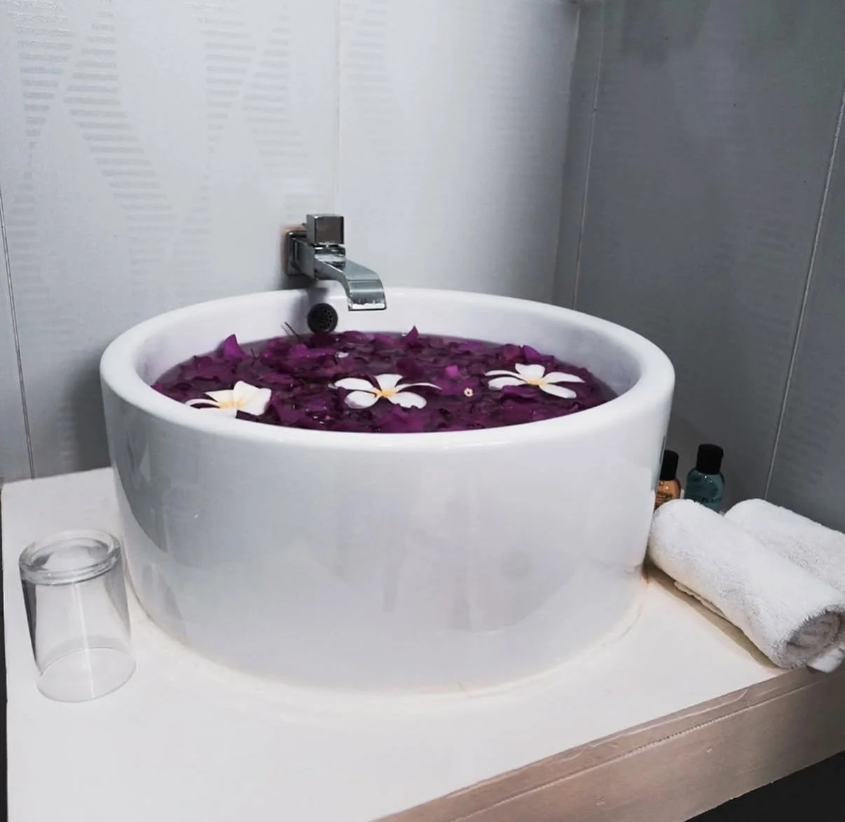 Lavabo rempli d'eau violette avec des fleurs blanches flottantes, barres d'ongles, serviette blanche, petits flacons de lotion et verre à côté.