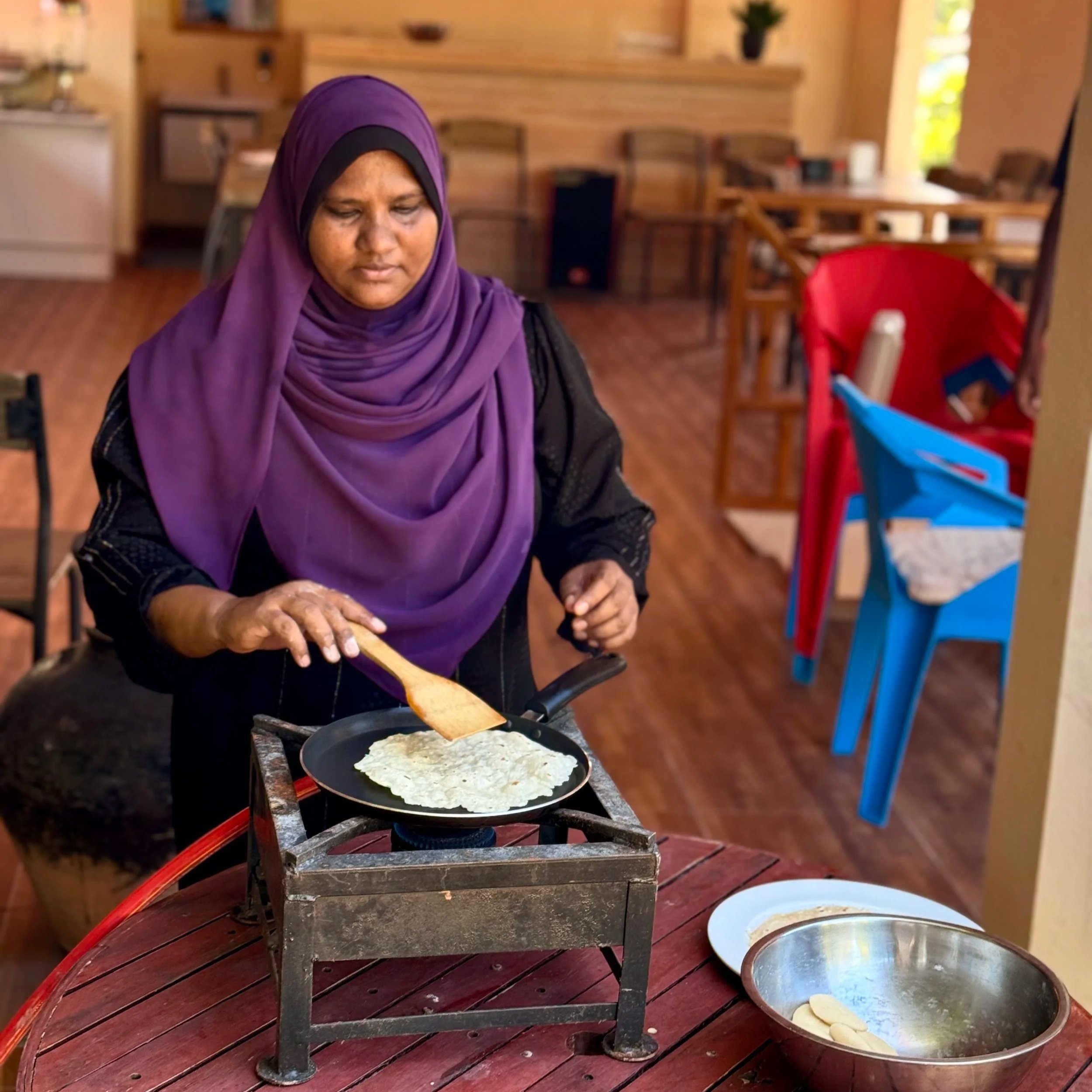Une femme portant un hijab violet et noir prépare une pâte à cuire sur une poêle sur un réchaud à gaz dans une cuisine rustique en bois.