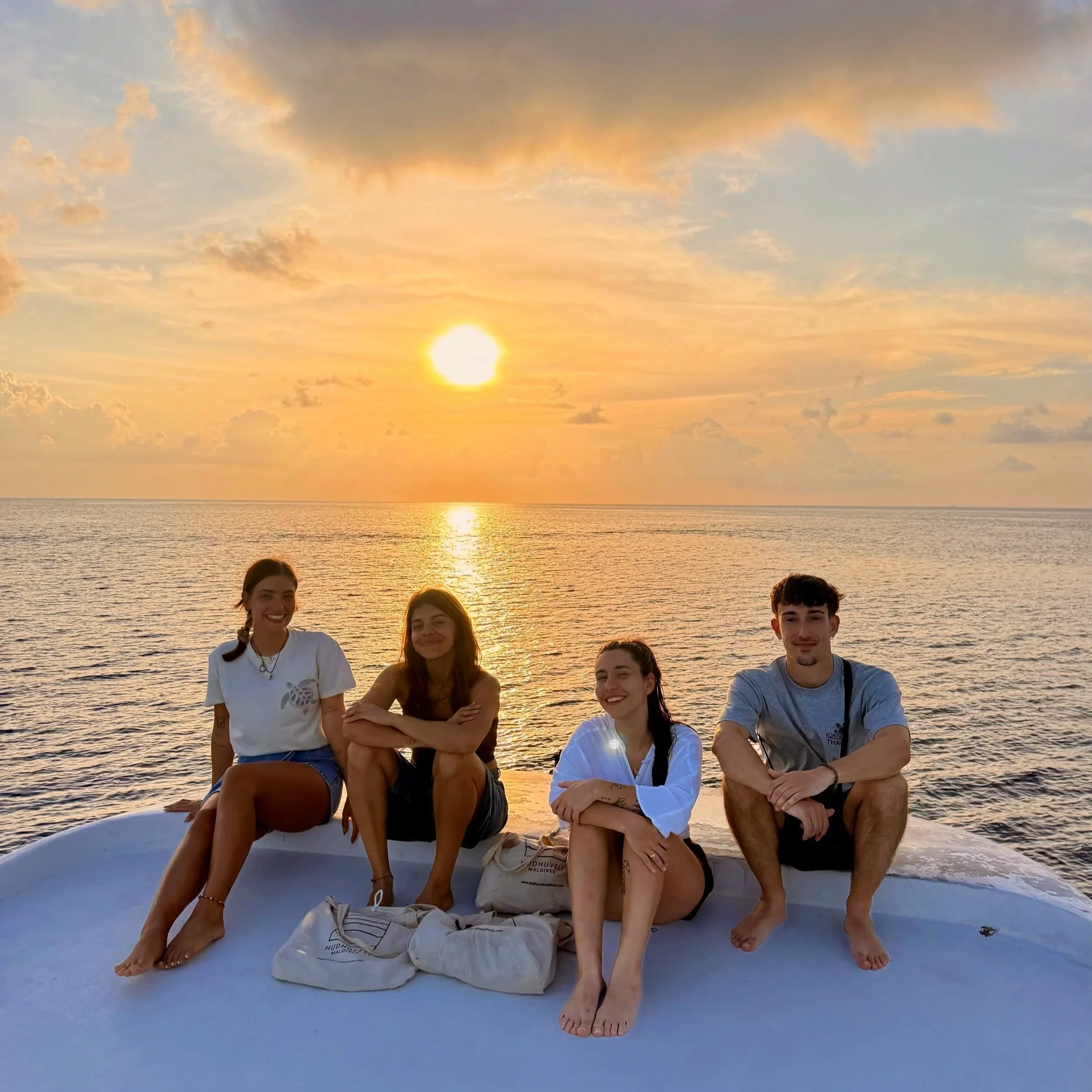 Groupe de jeunes assis sur un bateau au coucher du soleil à la mer, souriant et regardant vers l'objectif, avec des nuages colorés dans le ciel.