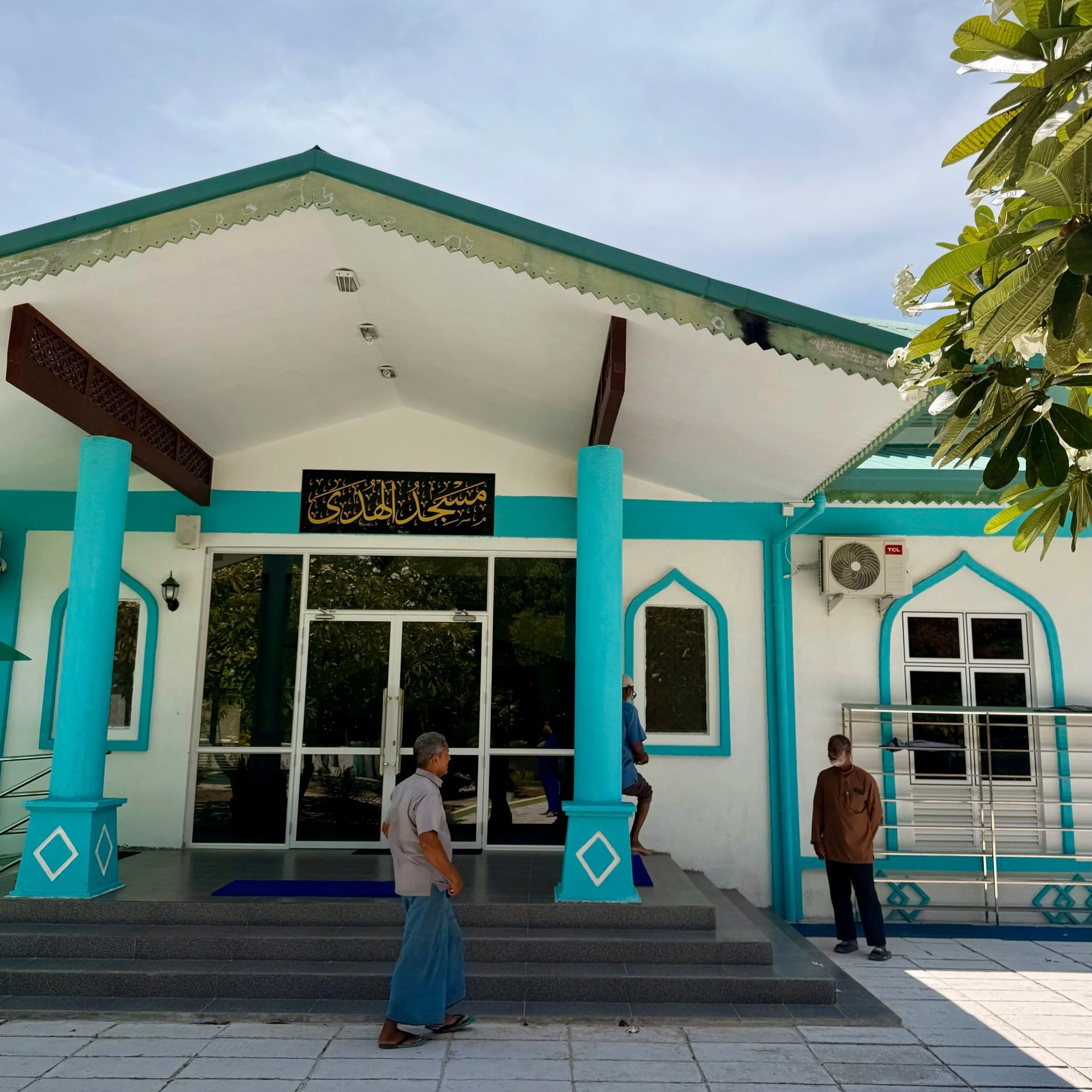 Façade d'un bâtiment avec un auvent turquoise, des colonnes bleues, des personnes debout devant, et un panneau avec de l'écriture arabe ou islamique au-dessus de la porte.