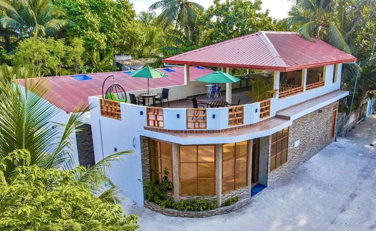 Une maison à deux étages avec terrasse, parasols, meubles et entourée de verdure tropicale.
