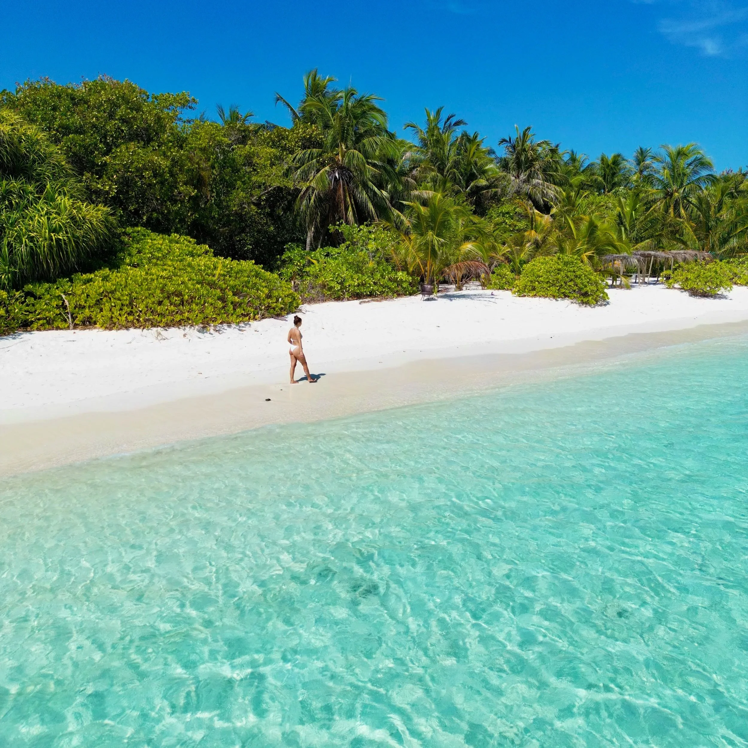 Une plage de sable blanc avec des eaux turquoise, une femme en maillot de bain marche au bord de l'eau, et une végétation tropicale avec des palmiers dense en arrière-plan.