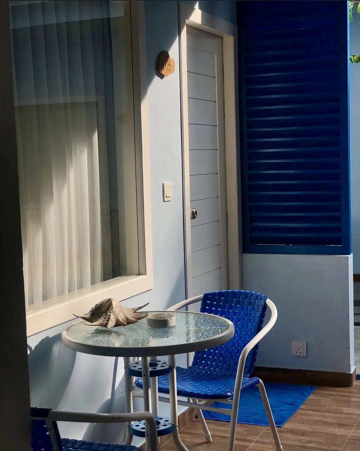 Petite terrasse avec une table en verre, deux chaises en plastique bleu, coquillage et cendrier, porte blanche, fenêtres avec volets bleus, sols en bois, mur bleu.