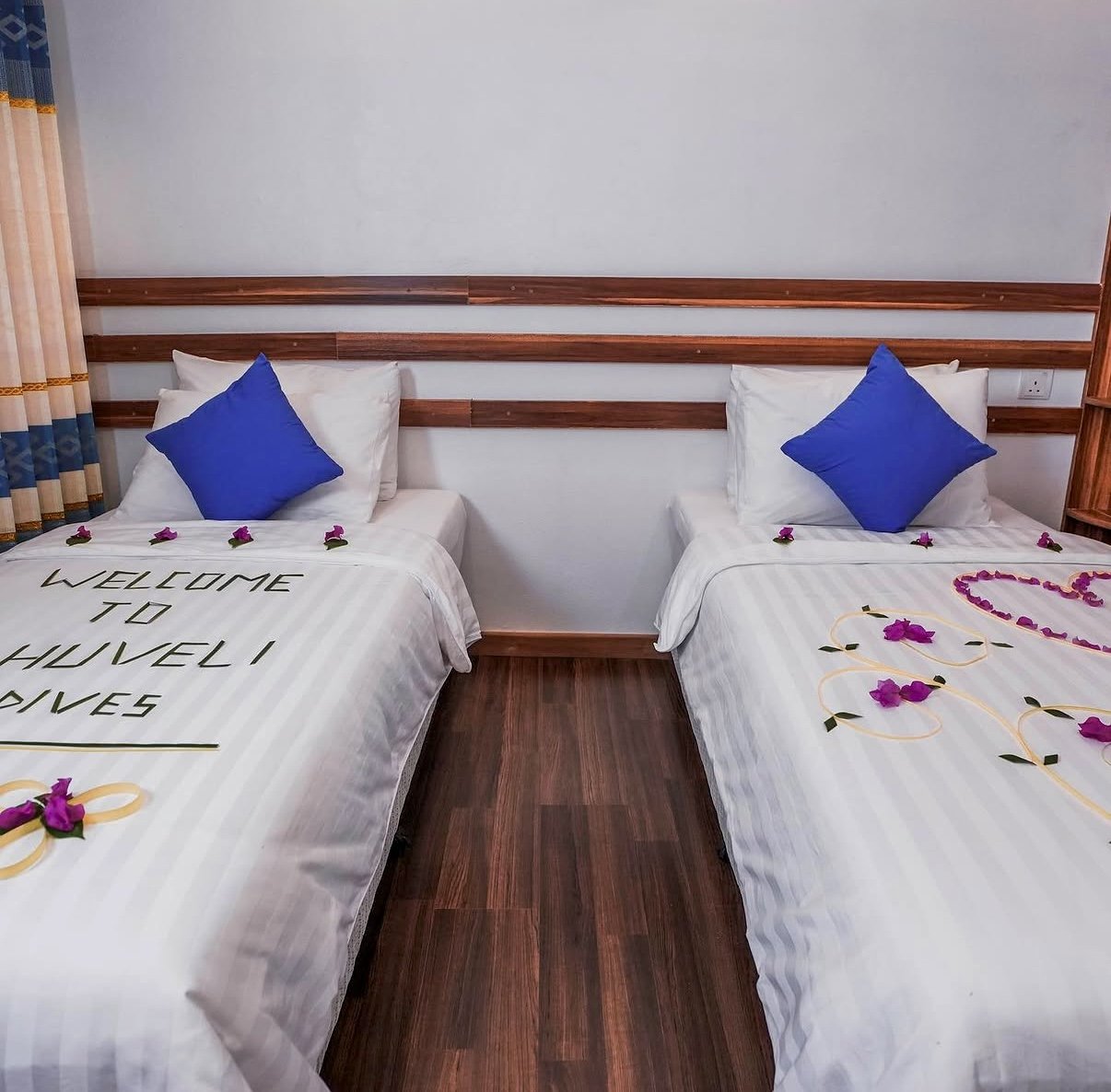 Deux lits dans une chambre d'hôtel décorés avec des pétales de fleurs et des cœurs en fleurs. L'un des lits a une écriture "WELCOME TO " sur la couverture, avec des oreillers blancs et un coussin bleu. Les lits sont séparés par un espace vide, avec u