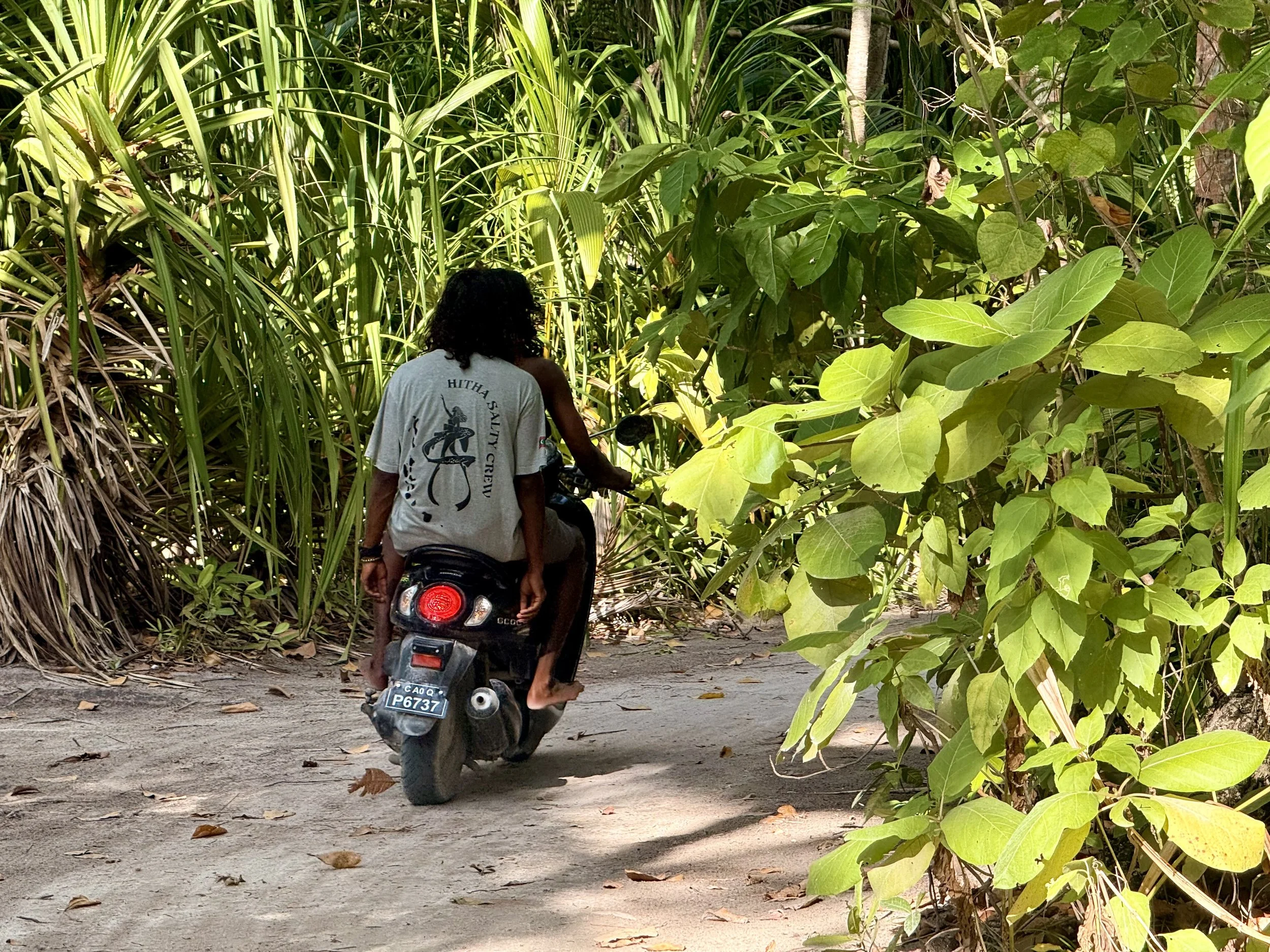 Une personne avec des cheveux bouclés portant un t-shirt portant une inscription en anglais est assise sur une moto verte, avançant sur un chemin entouré de végétation dense avec de grandes feuilles vertes.