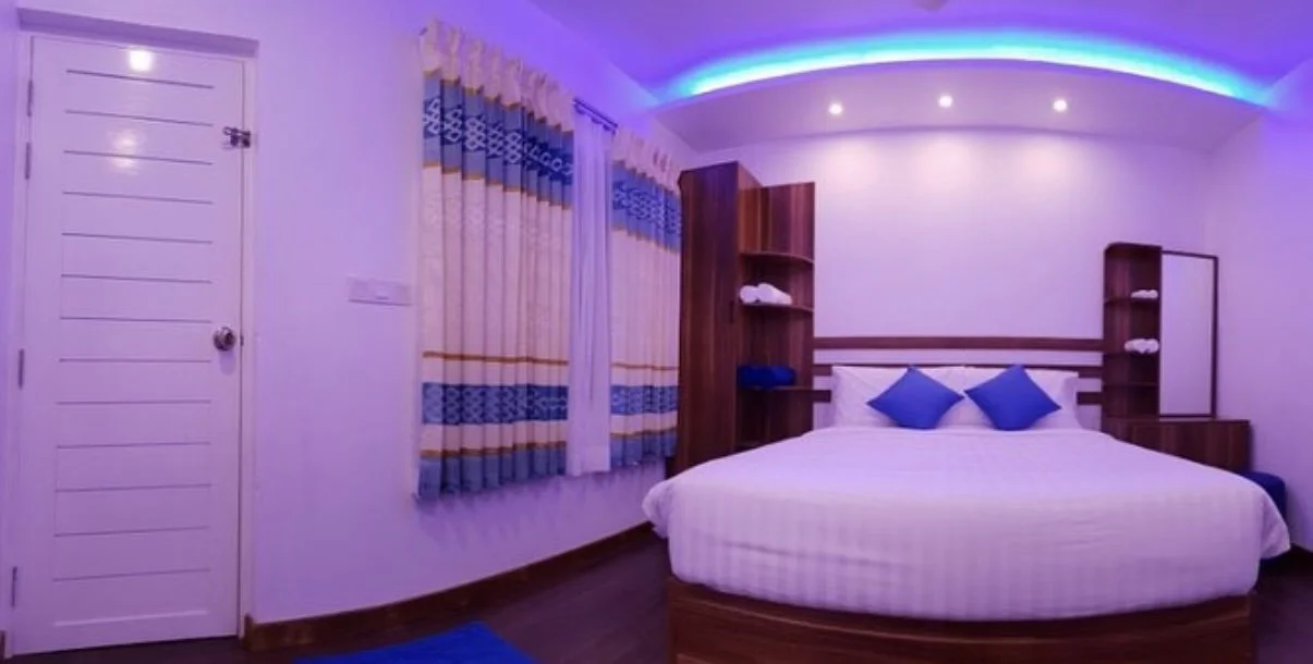 Une chambre avec un lit rond et deux coussins bleus, des étagères en bois, des rideaux à motifs bleu et beige, et un plafond avec éclairage LED violet.