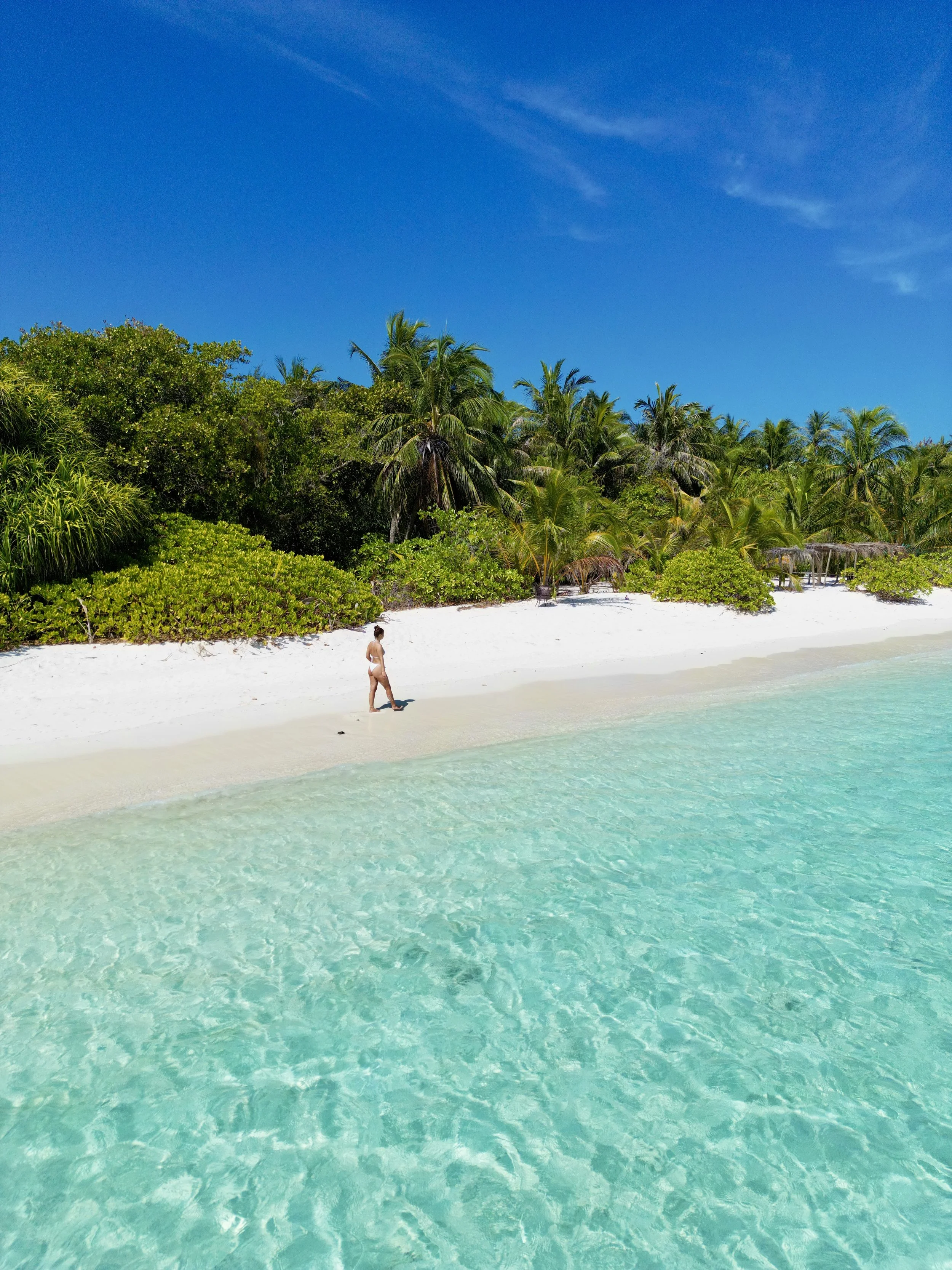 Plage de sable blanc avec une femme marchant près de l'eau turquoise, arrière-plan de végétation tropicale avec palmiers et ciel bleu clair.