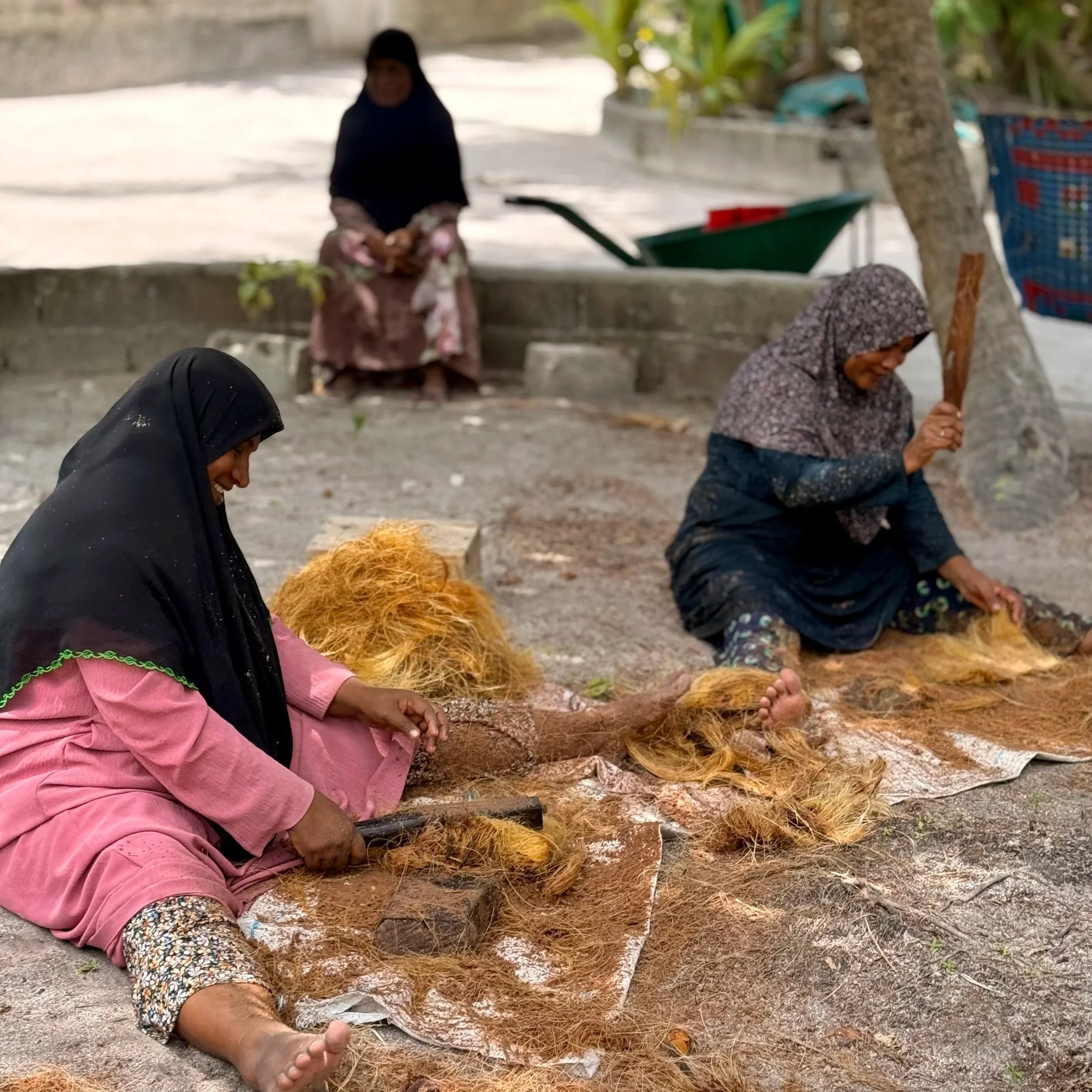 Deux femmes assises par terre en train de filer de la fibre de coco, avec un fond de végétation et un sol en béton.