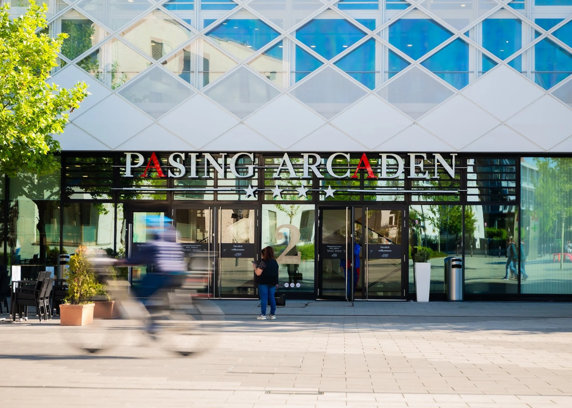 Bahnhofseingang mit Schild "Pasing Arcaden" und einigen Menschen davor, einige fahren vorbe, das Gebäude hat ein modernes Design mit Glasfassade