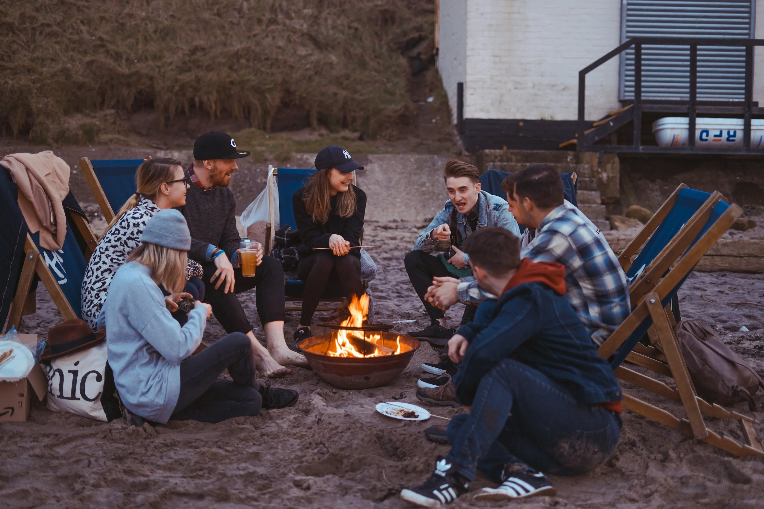 Group of friends sitting around a fire on the beach, enjoying conversation and drinks in the evening, with chairs and backpacks around them.
