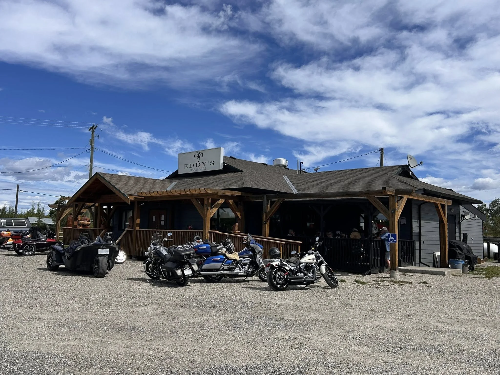 A gravel parking lot in front of a small restaurant named Eddy's Bar & Grill with several motorcycles parked outside, clear blue sky with scattered white clouds above.